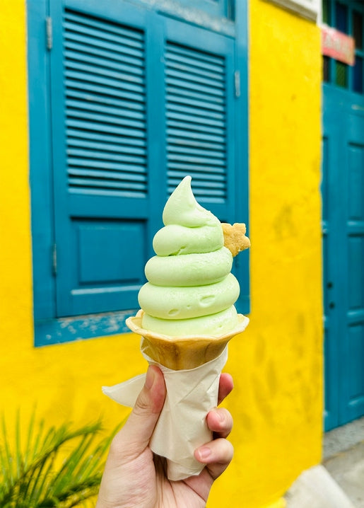 Hand holding pistachio ice cream cone against a yellow wall with blue shutters