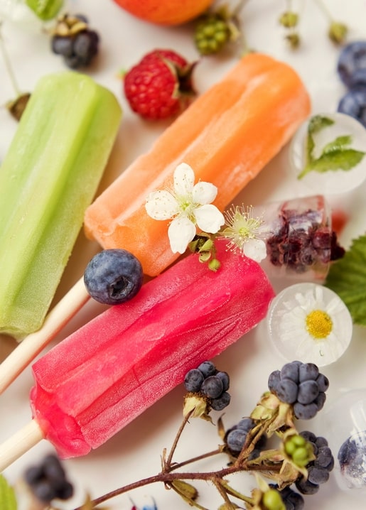 Grünes, orangefarbenes und pinkes Eis am Stiel zwischen Beeren, weißen Blüten und Eiswürfeln mit Beeren
