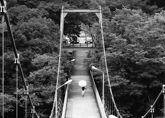 Person with a white umbrella walking across a suspension bridge over a forested valley