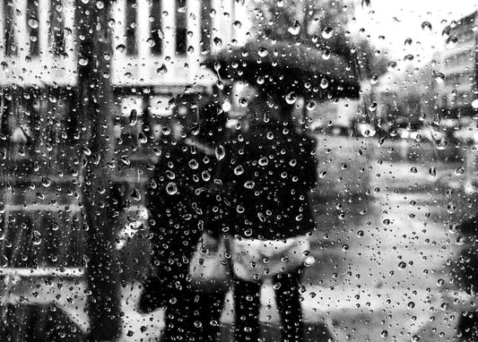 Black-and-white photo of raindrops on glass blurring a couple under an umbrella on a city street