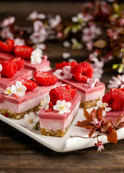 Raspberry cheesecake bars topped with fresh raspberries and cherry blossoms on a white plate