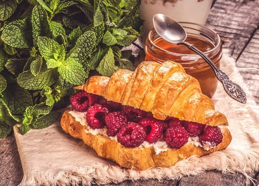 Croissant filled with cream and fresh raspberries beside a jar of honey and a bunch of mint