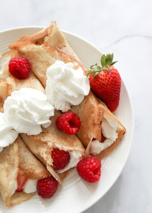 Gefaltete Crêpes mit Schlagsahne, Himbeeren und Erdbeere auf weißem Teller