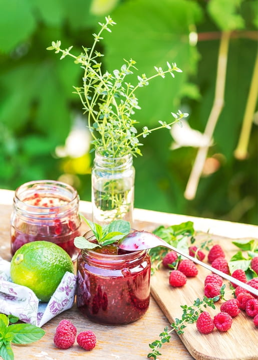Gläser mit Himbeermarmelade, frischen Himbeeren, Minze und Limette auf einem Gartentisch