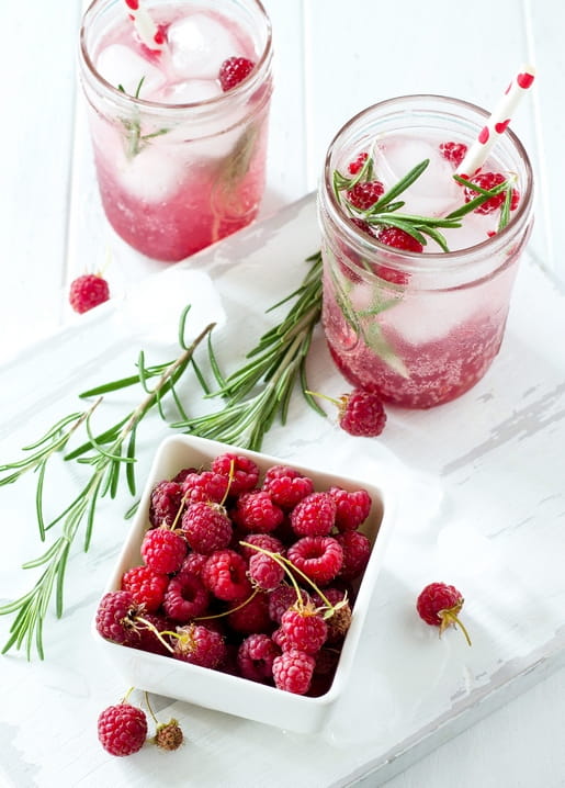 Zwei Gläser Himbeerlimonade mit Eis und Rosmarin neben einer Schale frischer Himbeeren