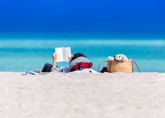 Person lying on white sand reading a book beside a woven beach basket and turquoise sea