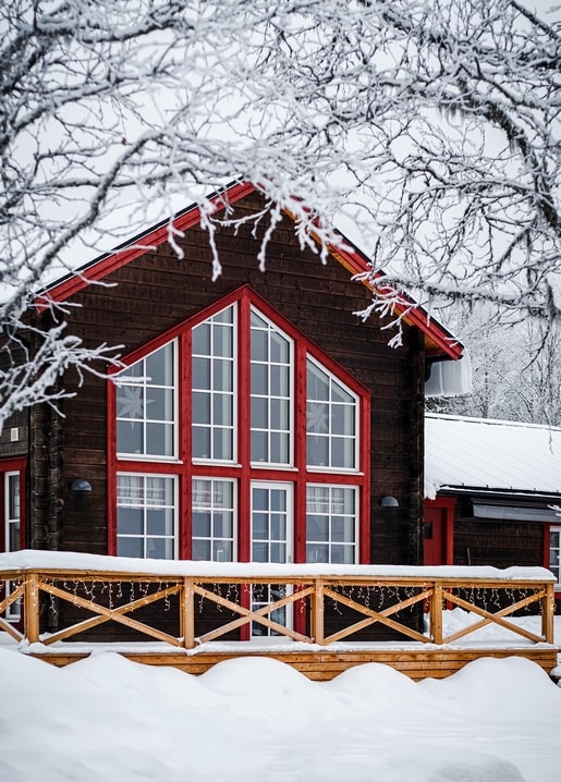 Dark wood cabin with red window frames behind snow-laced branches