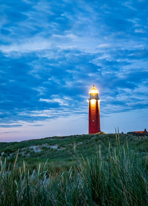 Red lighthouse with lit beacon glowing at dusk above dune grass
