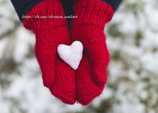 Zwei rote gestrickte Fäustlinge halten ein kleines weißes gehäkeltes Herz vor Schnee