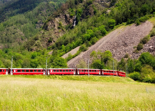 Roter Schmalspurzug fährt durch ein grünes Alpental an einem Wasserfall vorbei