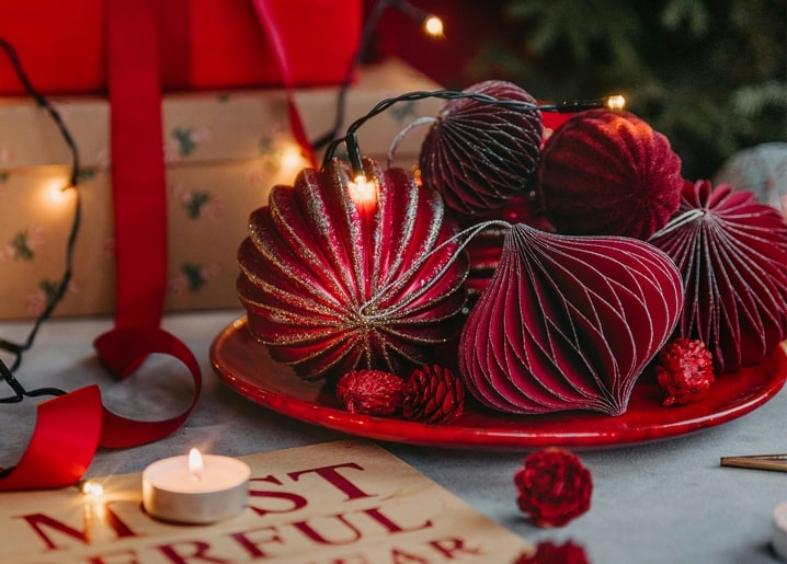 Red velvet and paper ornaments, pinecones, and a lit candle on a plate beside a wrapped gift and fairy lights