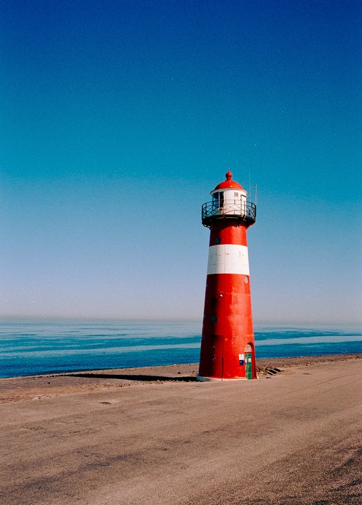 Red-and-white striped lighthouse against a clear blue sky, calm sea and empty beach in front.