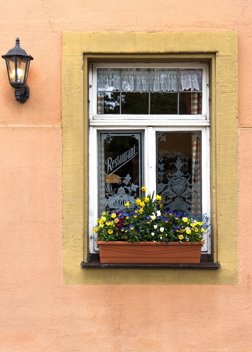 Weiß gerahmtes Restaurantfenster mit Spitzenvorhang, Laterne und Stiefmütterchen-Kasten