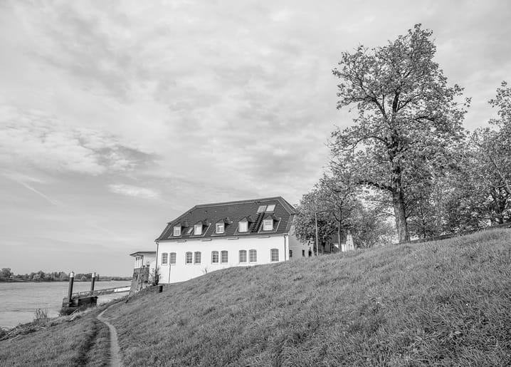 Black-and-white photo of a gabled house on a riverside dike beneath a large tree