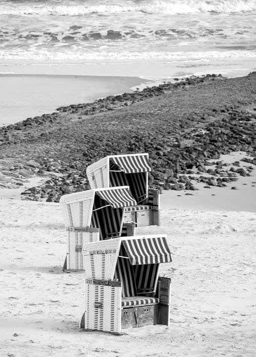 Three roofed wicker beach chairs lined up on sand with waves breaking along a rocky shoreline.