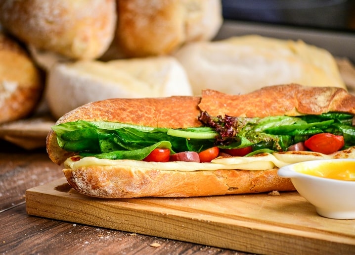 Sandwich mit Tomate und Salat auf Holzbrett, im Hintergrund frische Brötchen