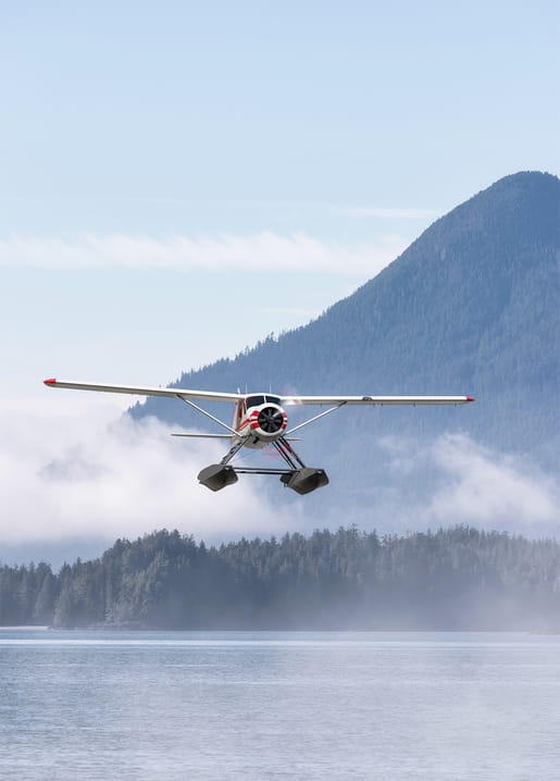 Small red-and-white seaplane flying low over a misty lake with a forested mountain behind it