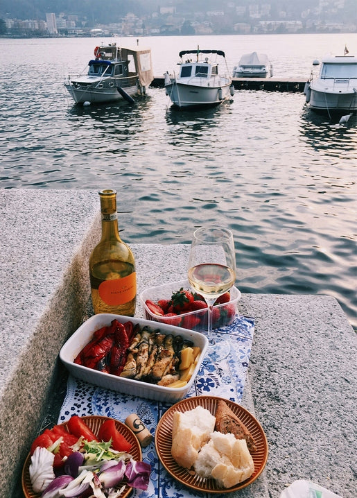 Seaside picnic spread of wine, strawberries, roasted mussels and bread with boats moored behind
