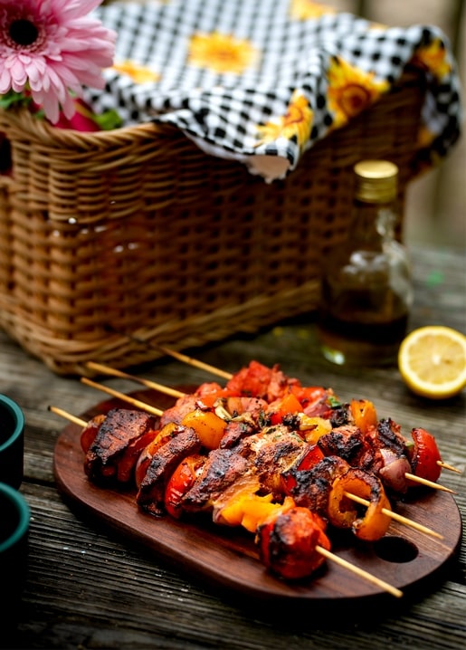 Grilled shashlik skewers on a wooden board beside a wicker picnic basket