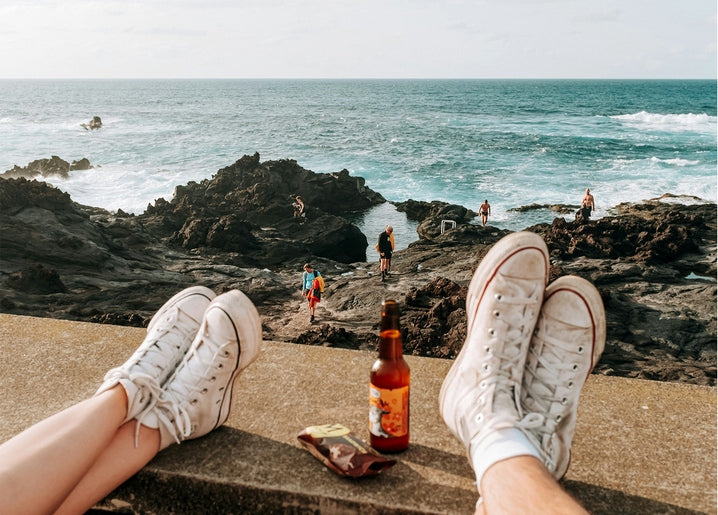 Zwei Paar Sneaker auf einer Mauer über felsiger Küste, dazwischen Bier und Snacks