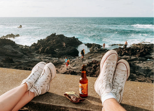 Two pairs of sneakers on a ledge above a rocky coastline, with beer and snacks between them