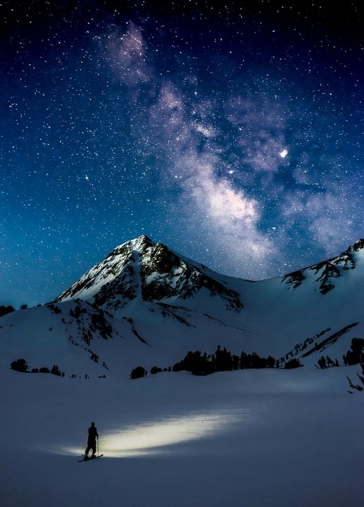 Silhouette of a snowboarder with a headlamp beneath a snow-capped peak and the Milky Way
