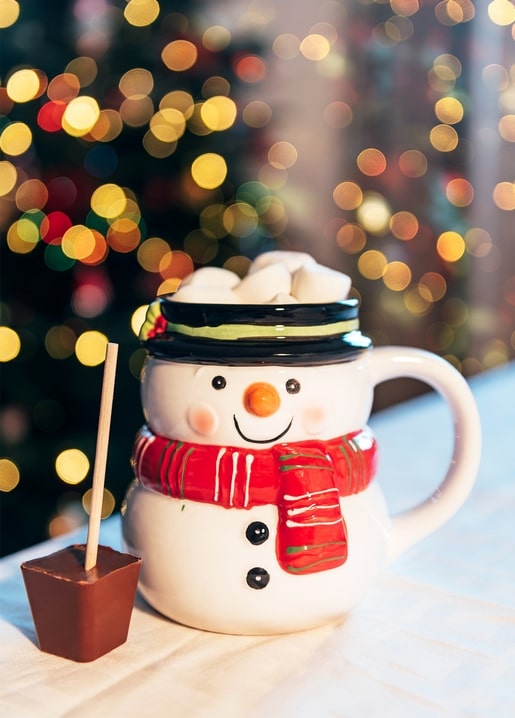 Snowman-shaped mug topped with marshmallows and a chocolate stick, Christmas lights behind