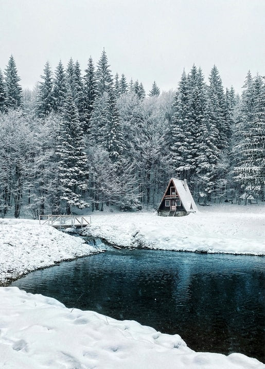 Snow-covered A-frame cabin beside a dark forest pond with a small footbridge