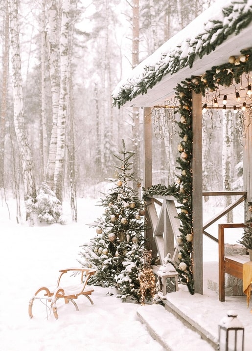 Snow-covered cabin porch decorated with pine garland, lights, a small tree and an old wooden sled