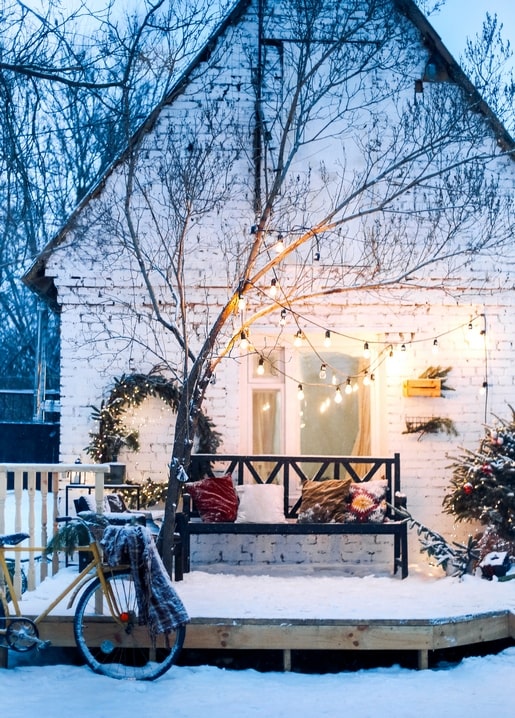 Snow-covered cottage porch with string lights, cushioned bench and a vintage bicycle at dusk