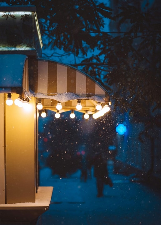 Snow falls at night on a striped market kiosk awning strung with warm fairy lights