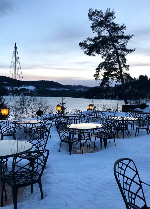 Snow-covered café terrace with empty chairs overlooking a frozen lake at dusk