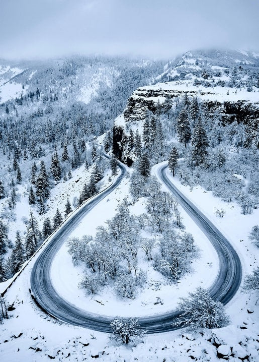 Verschneite Bergstraße mit enger Haarnadelkurve durch Nadelwald, von oben gesehen
