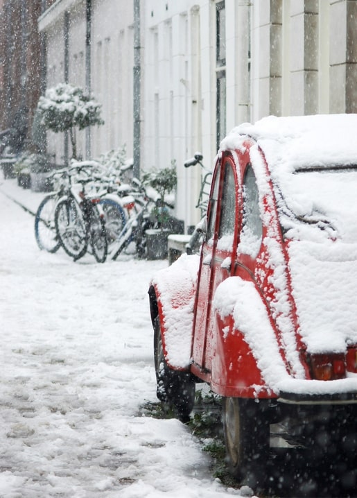 A vintage red car covered in snow parked beside bicycles on a European street during snowfall