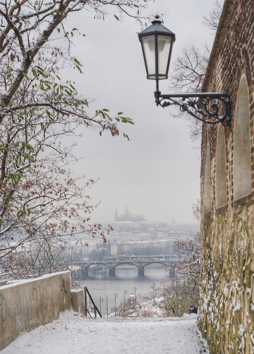 Snowy stone staircase and antique lamp overlooking Prague Castle across the Vltava