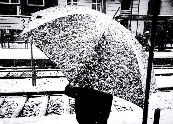 Person under a snow-covered umbrella walking across a train platform during a blizzard