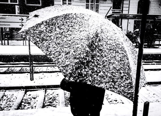 Person under a snow-covered umbrella walking across a train platform during a blizzard