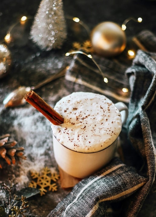 Frothy eggnog with a cinnamon stick beside gold Christmas baubles and warm fairy lights