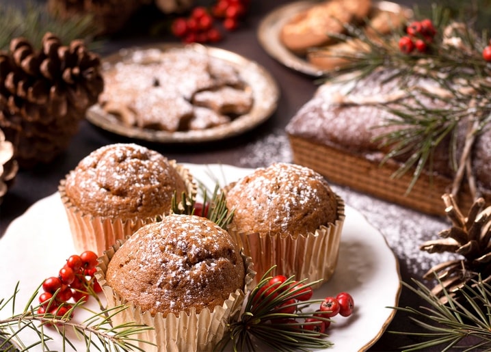 Powdered-sugar muffins on a plate with pine sprigs and red berries, holiday pastries blurred behind