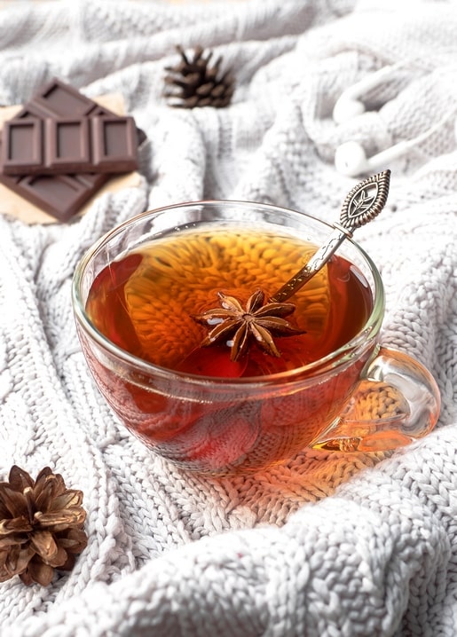 Glass cup of spiced tea with star anise beside chocolate and pinecones on a knit blanket