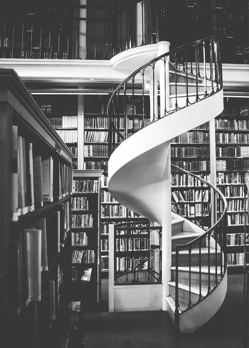 White spiral staircase winding upward between tall bookshelves in a black-and-white library photograph.