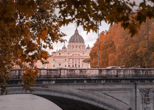 Blick auf die Kuppel des Petersdoms in Rom, umrahmt von Herbstlaub und einer Steinbrücke