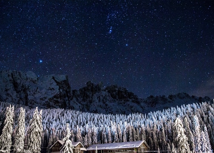 Starry night sky with Orion above a snow-covered pine forest and cabin roof