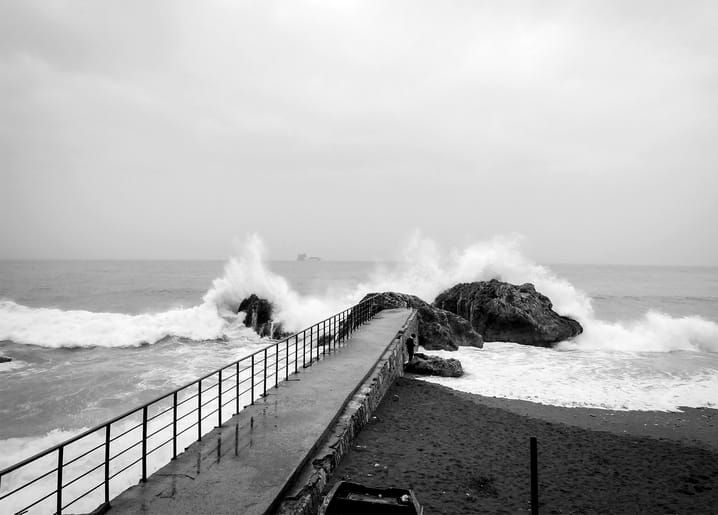 Storm waves crashing over rocks beside a concrete pier walkway with a metal railing