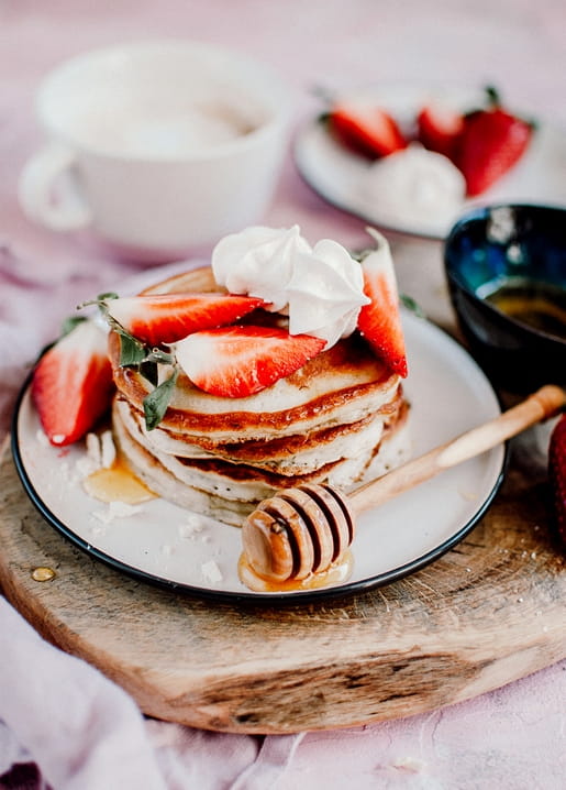Stack of pancakes topped with strawberries, whipped cream and honey drizzle on a wooden board