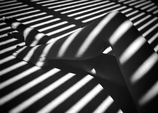 Black-and-white photo of striped sunlight and shadow across bare legs and feet