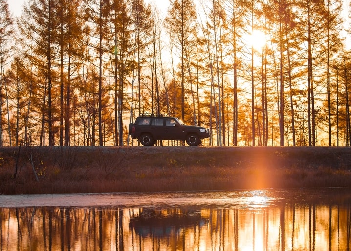 Dunkler Geländewagen fährt bei Sonnenuntergang an goldenen Lärchen vorbei, gespiegelt im See