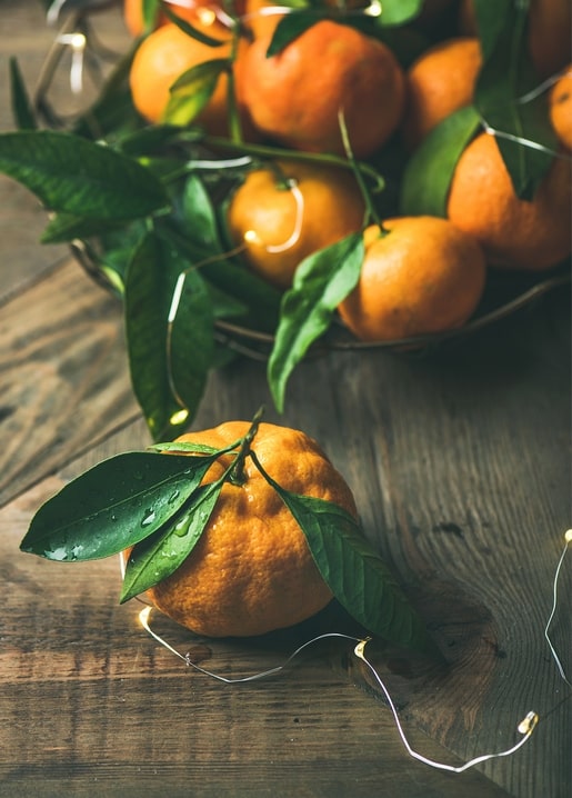 Fresh tangerine with green leaves and dew drops on a wooden table, basket of fruit behind