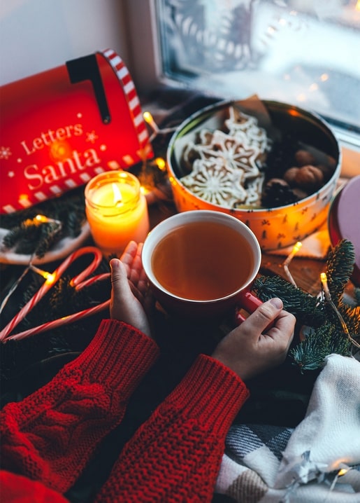 Hands in red mittens holding tea beside a Santa mailbox and candle