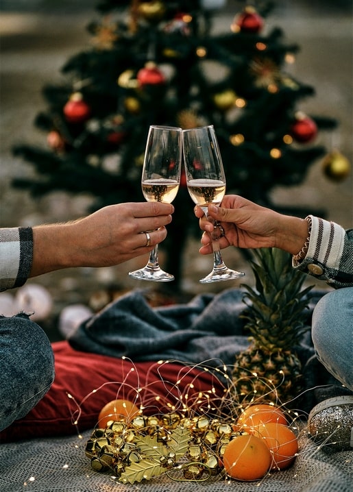 Two hands toasting champagne glasses in front of a Christmas tree with pineapple and oranges